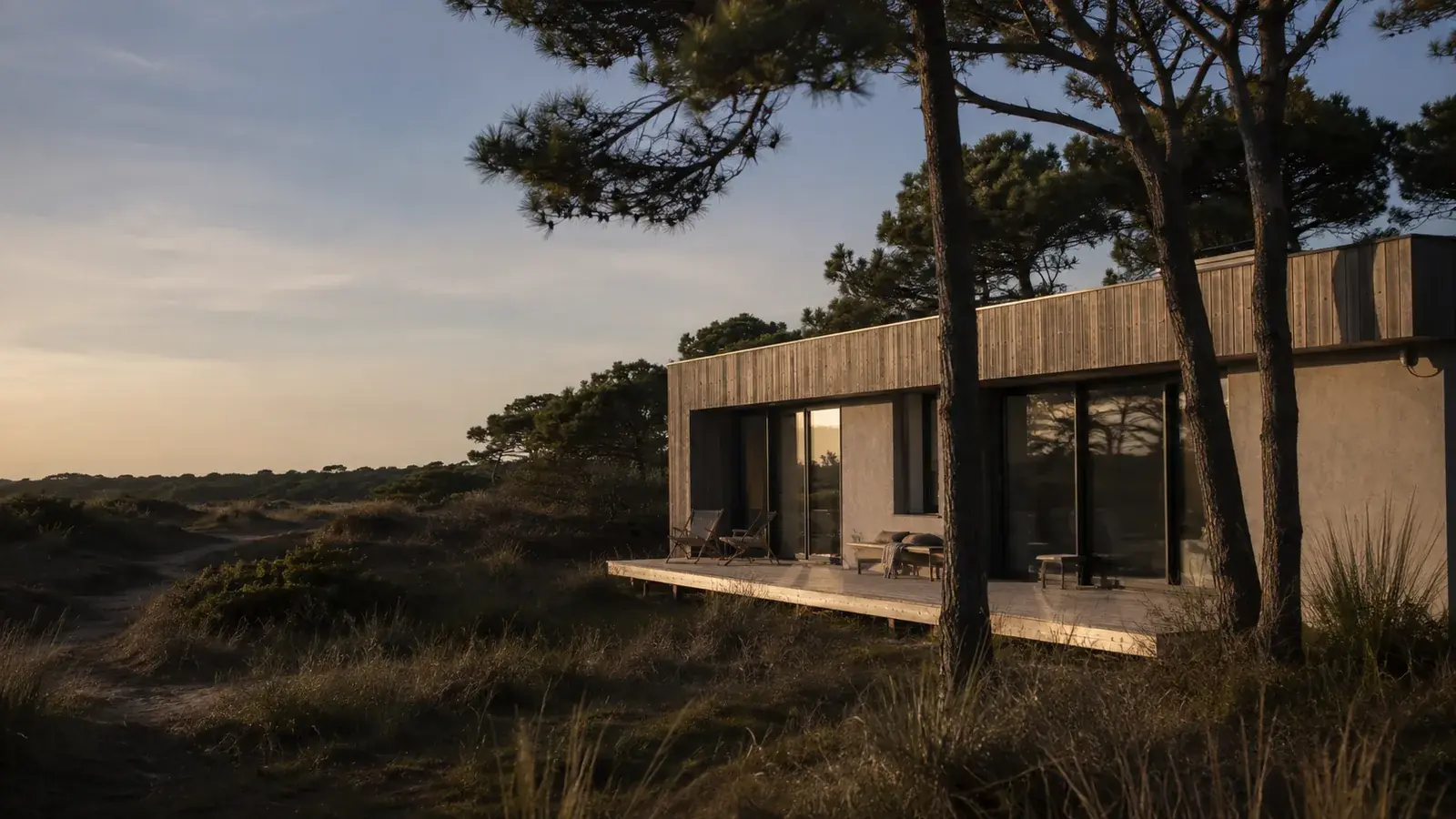 Maison fictive en bois et enduit clair, entourée de pins et de dunes
